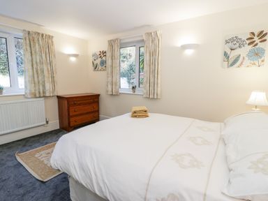 A bedroom with a bed covered in white linens a wooden dresser two windows with curtains and floral wall art at 11 Glyn Garth Mews in Llandegfan near Beaumaris