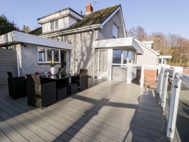 An outdoor deck area with a table, six chairs, a vase with flowers, and a house in the background at 11 Glyn Garth Mews in Llandegfan near Beaumaris