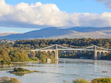 A suspension bridge over a river with hills and houses in the background at 11 Glyn Garth Mews Llandegfan near Beaumaris