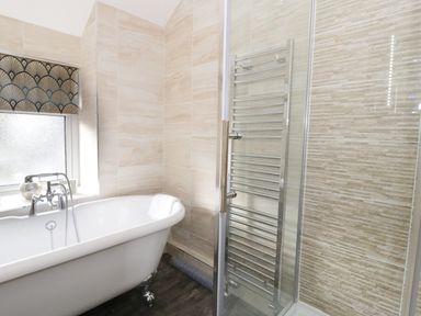 A bathroom with a white bathtub, a shower enclosure, a towel radiator, and a window with a patterned blind at Llwyn Onn in Dolwyddelan