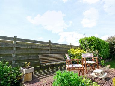 A wooden bench and two striped chairs on a wooden deck with plants and a wooden fence in the background at Blue Oyster in Mullion near Lizard Point