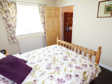 A bedroom with a wooden bed with floral bedding and purple pillows next to a window with patterned curtains at The Packing House in Hanley Castle near Malvern