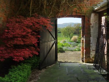 An open doorway in a brick wall with a black door leading to a garden with greenery and hills at The Packing House in Hanley Castle near Malvern