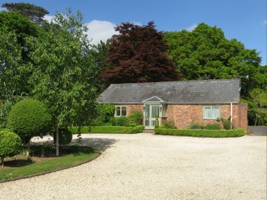 A small brick house with a gravel driveway and various green trees and bushes at The Packing House Hanley Castle near Malvern