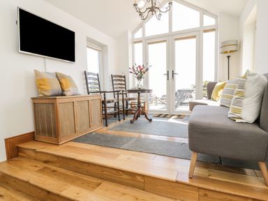 A sitting area with a gray sofa with striped cushions wooden chairs a small round table with flowers and a wall-mounted television at Marsh Villa in Silloth