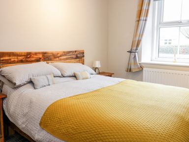 A bedroom with a bed with yellow blanket pillows a wooden headboard bedside tables and a window with curtains at Victoria Cottage in Norham