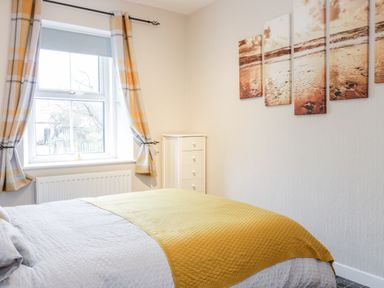 A bedroom with a bed covered by a white and yellow blanket next to a window with yellow plaid curtains and a white chest of drawers at Victoria Cottage in Norham