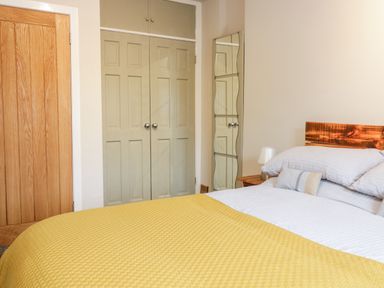 A bedroom with a bed with yellow blanket and white pillows next to a wooden door and green closet doors with a wall mirror at Victoria Cottage in Norham