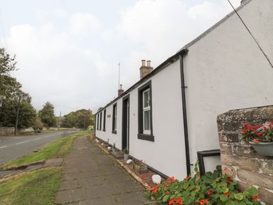 A white cottage with black window frames and a paved sidewalk in front near a road with grass and trees at Victoria Cottage in Norham