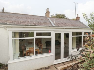 Exterior view of a single-story house with large windows and a double glass door showing a dining table and chairs inside at Victoria Cottage in Norham