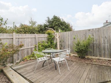 A wooden deck with a round glass table and four metal chairs in a fenced garden at Victoria Cottage in Norham