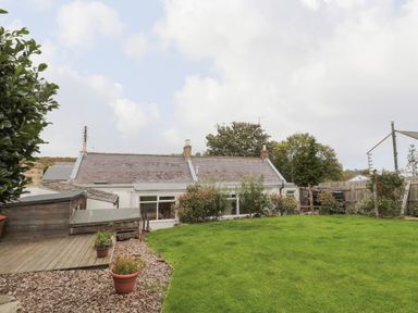 A backyard garden with a grassy lawn wooden decking and a white house with a slate roof at Victoria Cottage in Norham