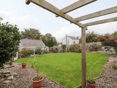 A garden with green grass surrounded by plants and a wooden pergola with houses visible in the background at Victoria Cottage in Norham