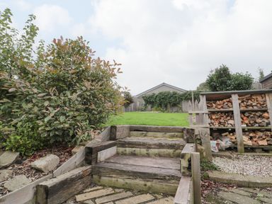 Wooden steps leading to a grassy area with stacked firewood and plants at Victoria Cottage in Norham