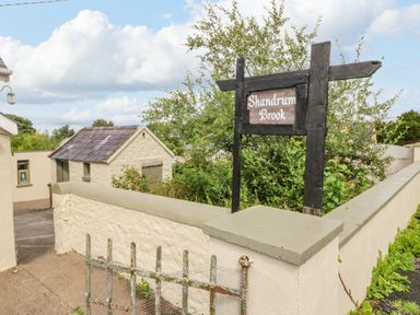 An exterior view of a building with a small shed a stone wall and a wooden sign reading Shandrum Brook in Charleville