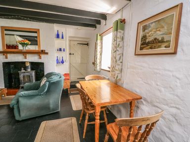 A dining table with wooden chairs next to a window and armchair near a fireplace with a mirror and blue bottles on shelves at Taliharris Cottage in Llandysul