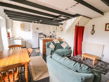 A living area with two sofas a wooden table and chairs a small wooden coffee table a kitchen with appliances and a staircase with a red curtain at Taliharris Cottage in Llandysul