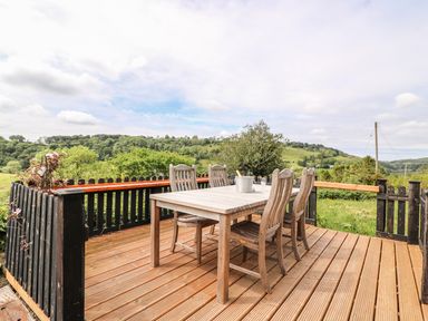 An outdoor wooden deck with a table and six chairs overlooking green hills at Taliharris Cottage in Llandysul
