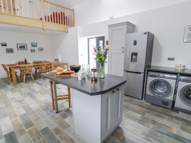 A kitchen with an island counter set with food and flowers a dining table with chairs and a washing machine and dryer at Sgubor in Pentraeth