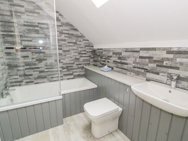 A bathroom with a bathtub shower combination toilet and sink with gray tiled walls and wood paneling at Sgubor in Pentraeth
