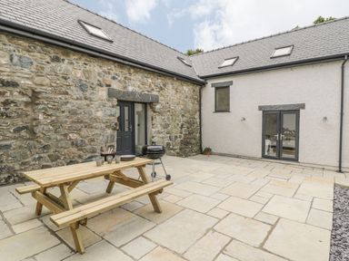 An outdoor patio area with a wooden picnic table and grill outside stone and white buildings at Sgubor in Pentraeth