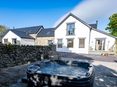 A hot tub near a stone wall and a house with multiple windows and doors at Sgubor in Pentraeth