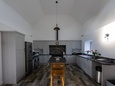A kitchen with grey cabinets a black countertop a wooden chair an oven under a brick backsplash a refrigerator and a washing machine at Sgubor in Pentraeth