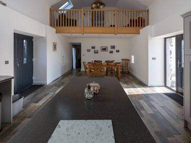An open kitchen and dining area with a wooden railing above and a dining table set with wine glasses at Sgubor in Pentraeth