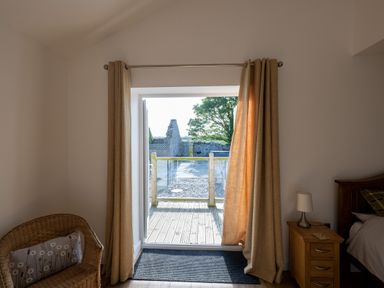 A room with glass door and beige curtains opening to a wooden deck and outside view at Sgubor in Pentraeth