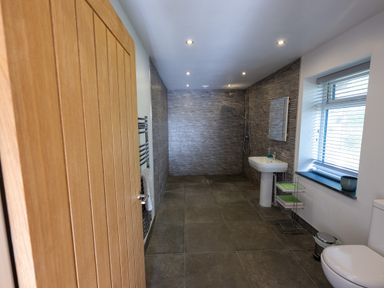 A bathroom with a wooden door, sink, toilet, window with blinds, towel rack, and a walk-in shower at Sgubor in Pentraeth