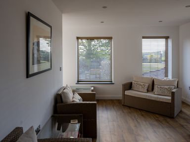 A sitting area with wicker armchairs and a sofa with cushions near windows with blinds at Sgubor in Pentraeth
