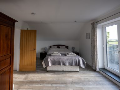 A bedroom with a bed with folded towels bedside tables lamps a wooden cabinet an open door and a window with floral curtains at Sgubor in Pentraeth
