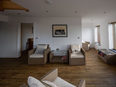 A sitting area with wicker chairs and a glass coffee table on wooden flooring with white walls and windows at Sgubor in Pentraeth