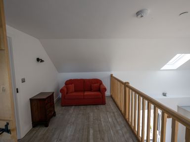 A red sofa with two cushions next to a wooden chest of drawers and a wooden railing under a sloped ceiling at Sgubor in Pentraeth