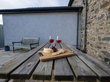 A wooden picnic table with cheese grapes crackers and two glasses of red wine outside next to a stone wall and bench at Sgubor in Pentraeth