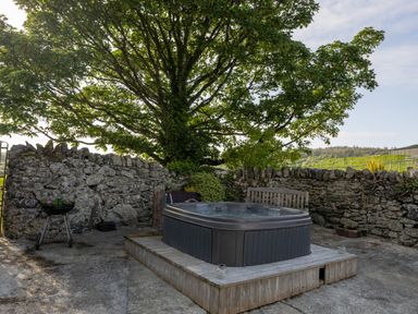 An outdoor area with a hot tub on a wooden platform under a large tree surrounded by a stone wall at Sgubor in Pentraeth