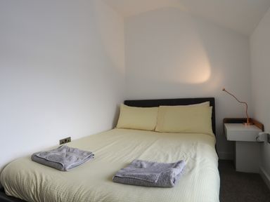 A bed with light yellow bedding and gray towels on it next to a white wall and a small white bedside table with a copper-colored lamp at The Old Post Office 23 High Street in Cemaes Bay
