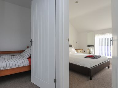 Two bedrooms separated by an open white door with one bedroom having a wooden bed and striped bedding and the other bedroom having a larger bed with white bedding a bedside table and a tall mirror at The Old Post Office 23 High Street Cemaes Bay