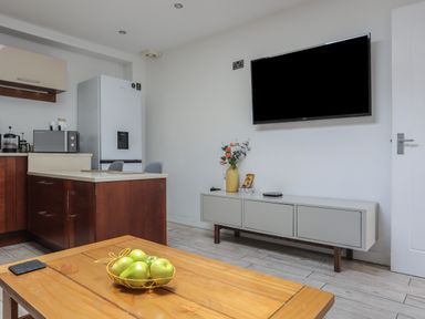 A kitchen and living area with a wooden table holding green apples a white cabinet with a vase and a TV mounted on the wall at The Studio @ The Old Post Office in Cemaes Bay