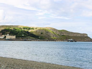 A rocky shoreline with water leading to a pier and hills with buildings at Little Orme View in Llandudno