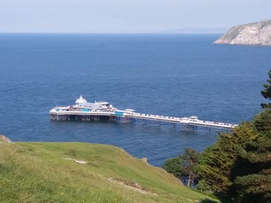A long pier extending into the sea with a building at the end and grassy hillside with trees along the shore at Little Orme View in Llandudno