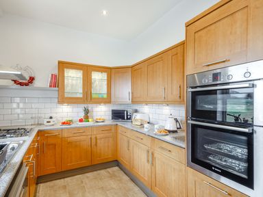 A kitchen with wooden cabinets, a double oven, microwave, stove, kettle, toaster, and countertop with fruit and mugs at Little Orme View in Llandudno