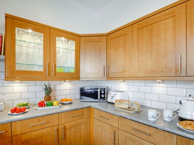 A kitchen corner with wooden cabinets a microwave toaster bread cups fruit and vegetables at Little Orme View in Llandudno