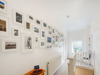 A hallway with framed pictures on the wall a small table with items a radiator a chair by a window at Little Orme View in Llandudno