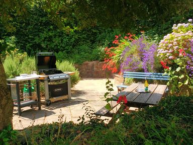An outdoor patio area with a barbecue grill a wooden picnic table with a wine bottle and glasses and colorful flowers surrounding the space at Beulah Cottage Green Bottom near Littledean