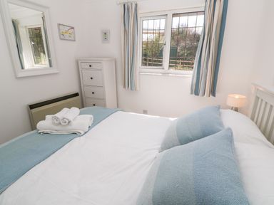 A bedroom with a double bed with blue pillows and folded towels on it a white chest of drawers a window with striped curtains and a bedside lamp at Rockpool Harlyn Bay near St Merryn