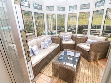 A sunroom with wicker sofa and chairs with cushions and a glass top table at Rockpool in Harlyn Bay near St Merryn