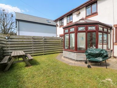 A garden with a wooden picnic table a barbecue covered in a green tarp and a conservatory attached to the house at Rockpool in Harlyn Bay near St Merryn