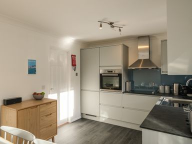 A kitchen with white cabinets a built in oven a stainless steel extractor fan a wooden sideboard with a fruit bowl and a speaker at Rockpool in Harlyn Bay near St Merryn