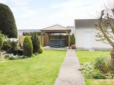 A backyard with a grassy lawn a concrete pathway a wooden gazebo with a hot tub and surrounding trees and plants at The Elms in Milford Haven